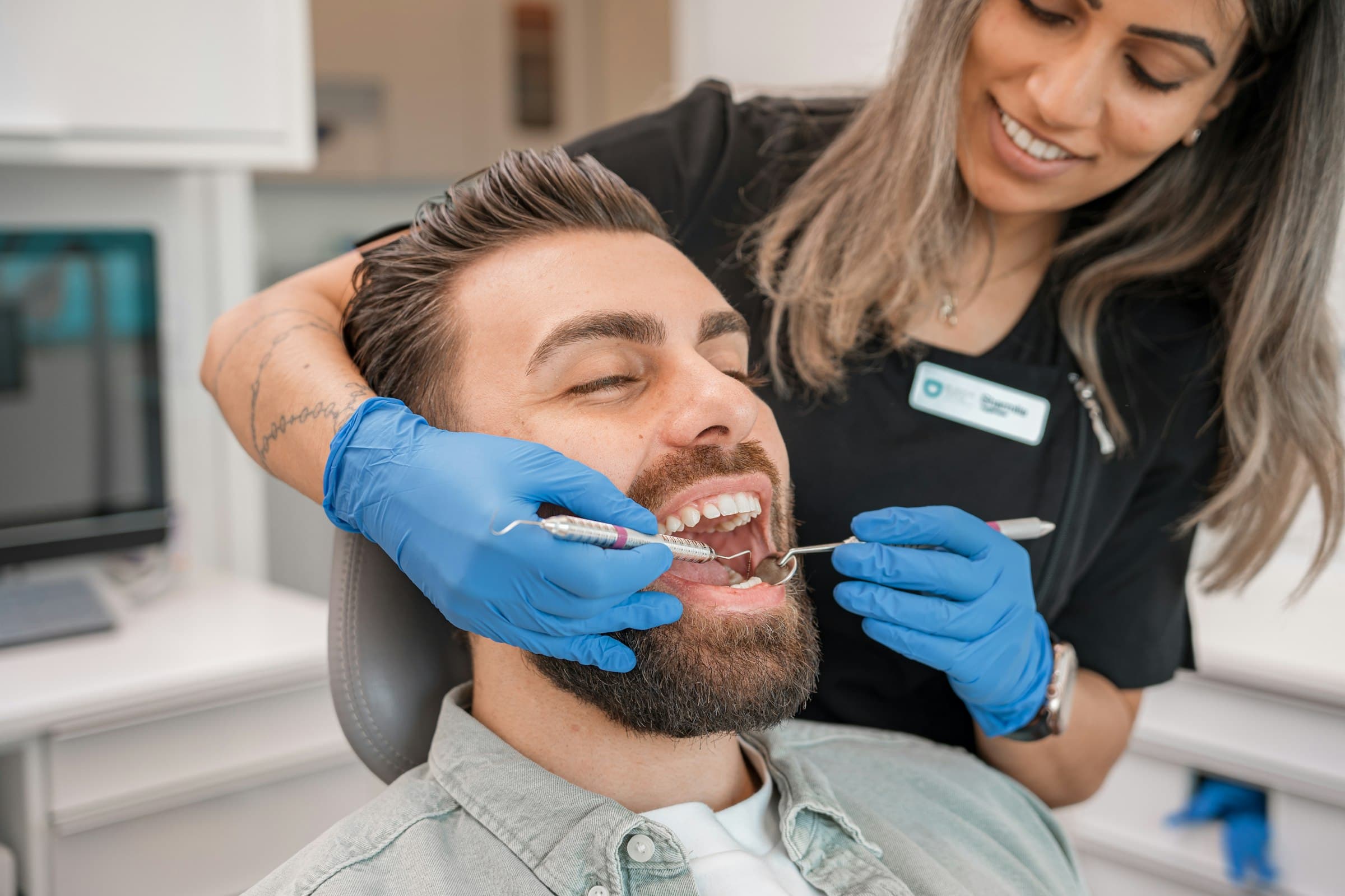 Paciente sorrindo na cadeira odontológica enquanto a dentista, de luvas azuis, examina seus dentes com instrumentos